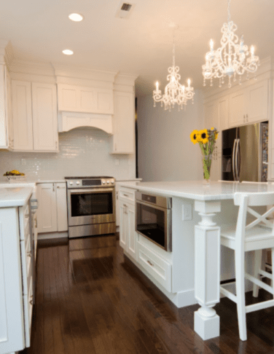 Modern kitchen interior featuring white cabinetry, stainless steel appliances, a central island with bar stools, and a crystal chandelier.