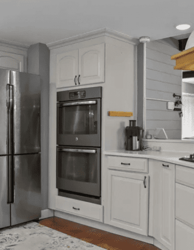 A modern kitchen with white cabinets, a stainless steel refrigerator, double wall ovens, a gas stovetop, and a window to the living room. The space is lit by recessed and hanging lighting.