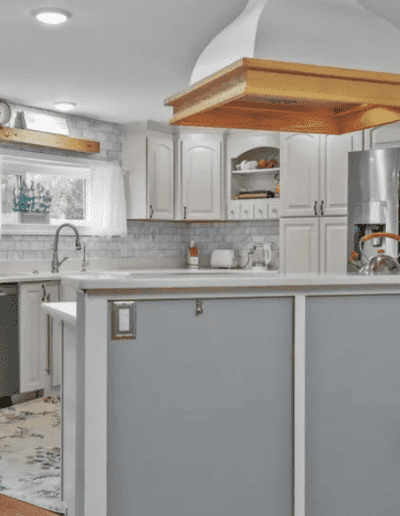 A modern kitchen with white and gray decor, featuring a central island with a cooktop, white cabinetry, stainless steel appliances, and wooden accents on the range hood and cabinetry.