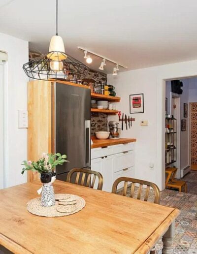 A cozy kitchen with a wooden dining table, metal chairs, a stainless steel fridge, open wooden shelves, and a view of a partially stone wall. A hallway with patterned tiles is in the background.