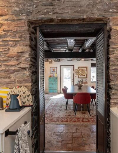 View through an open doorway in a rustic kitchen with stone walls, revealing a dining area with a table and red chairs, and beyond that a room with a turquoise dresser and patterned rug.