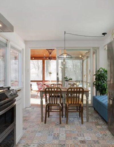 Modern kitchen with a gas stove, wooden cabinets, tiled floor, stainless steel refrigerator, and a dining area with a table and wooden chairs. Large windows provide natural light and a view outside.